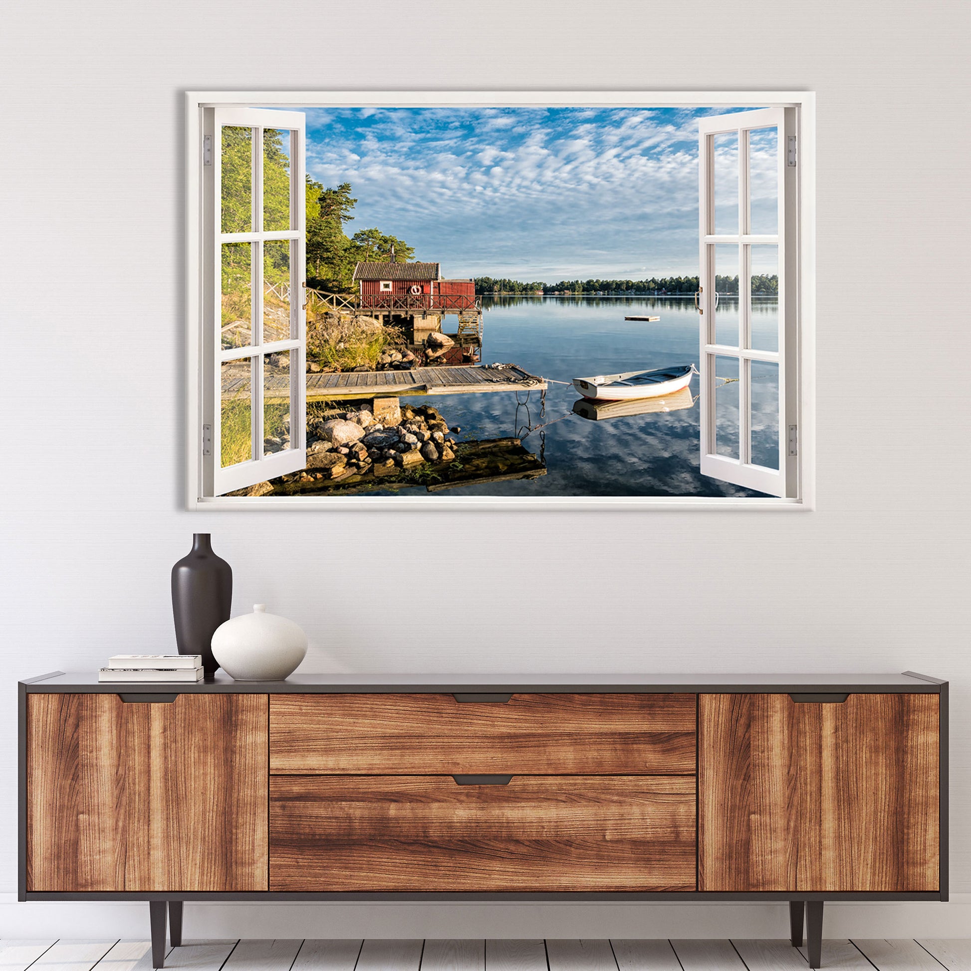 Ein Wandtattoo mit einer offenen Fensteransicht auf eine idyllische See-Landschaft, angebracht über einer modernen Holzkommode im Wohnzimmer. Der Blick führt auf einen kleinen Steg mit einem vertäuten Ruderboot, das sich auf dem ruhigen Wasser spiegelt. Links befindet sich ein rustikales, rotes Bootshaus auf Holzpfählen, umgeben von grünen Bäumen und einer felsigen Uferlandschaft. Der Himmel ist mit sanften Wolken bedeckt, die sich malerisch im Wasser spiegeln. Dieses Wandbild schafft entspannte Stimmung.