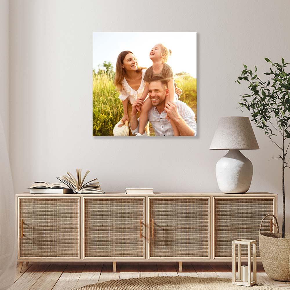 Stilvoll eingerichtetes Wohnzimmer mit einer natürlichen Holzkommode, dekorativen Büchern, einer modernen Tischlampe und einer grünen Pflanze. An der Wand hängt eine Fotoleinwand mit einem Familienbild, das eine glückliche Familie in der Natur bei Sonnenuntergang zeigt. Die warme Atmosphäre des Bildes ergänzt das gemütliche Wohnambiente.