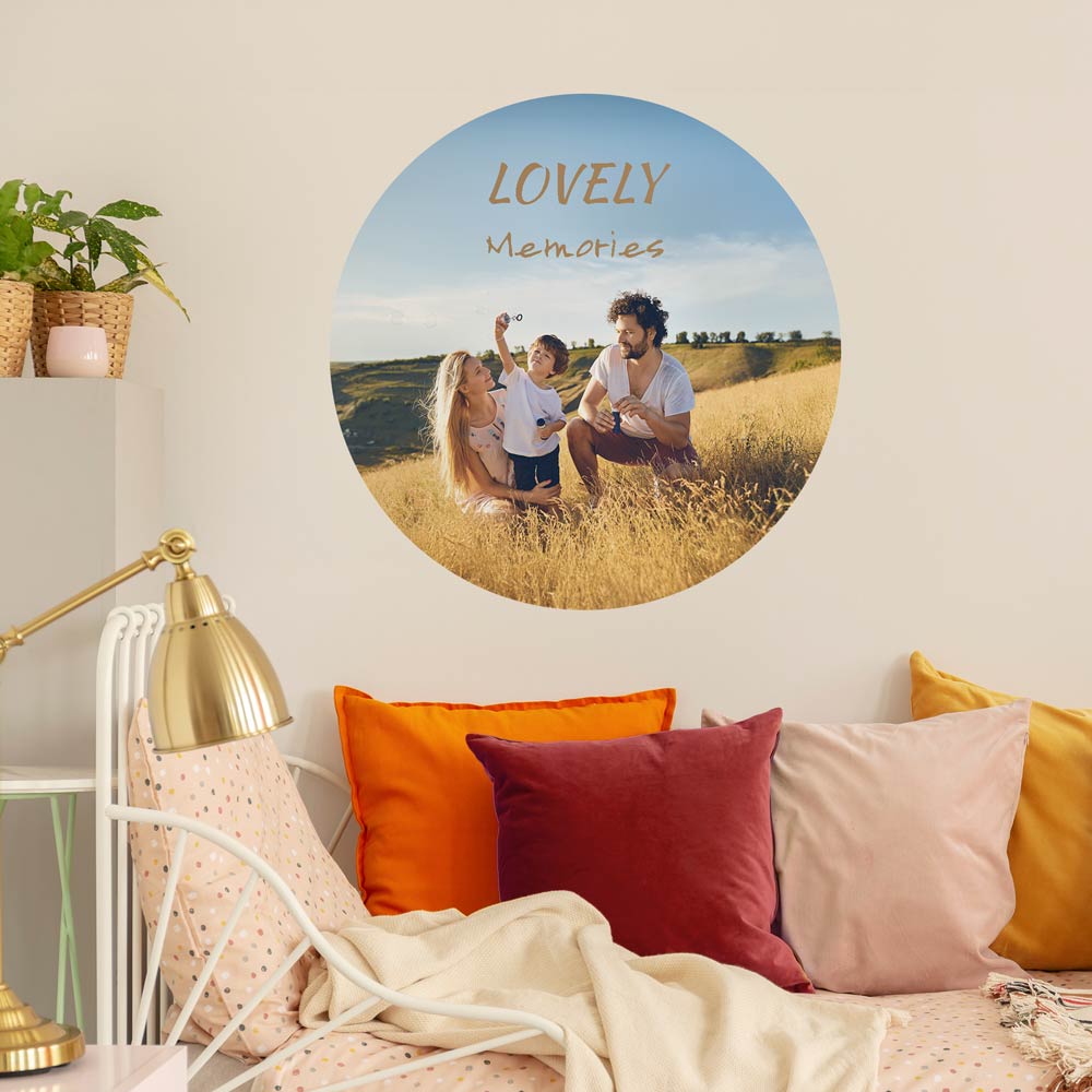 Großer, runder Wandsticker mit dem Schriftzug 'Lovely Memories' und einem Familienfoto auf einer Wiese. Der Sticker ist an der Wand eines gemütlich eingerichteten Schlafzimmers angebracht, dekoriert mit bunten Kissen, einer goldenen Lampe und Pflanzen. Perfekt für eine individuelle und persönliche Wandgestaltung.