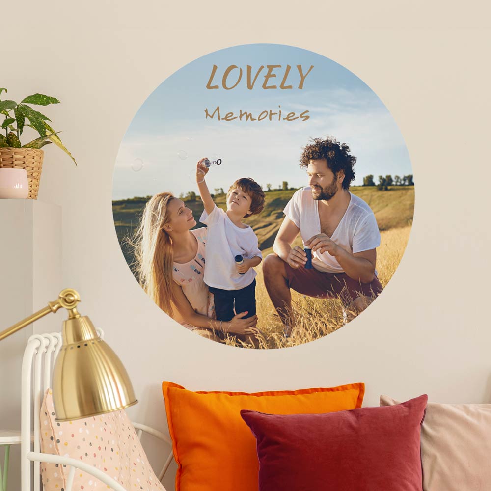 Runder Wandsticker mit dem Schriftzug 'Lovely Memories' und einem Familienfoto auf einer Wiese. Der Sticker ist an einer Wand in einem gemütlichen Wohnzimmer angebracht, dekoriert mit bunten Kissen, einer goldenen Lampe und einer Pflanze. Perfekt für individuelle Wandgestaltung und persönliche Erinnerungen.