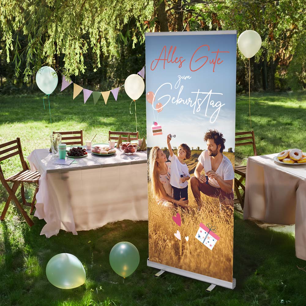 Outdoor-Geburtstagsfeier mit festlich dekorierten Tischen, Luftballons und einer Girlande im Grünen. Im Vordergrund steht ein personalisierbares Rollup-Banner mit dem Text 'Alles Gute zum Geburtstag' und einem Familienfoto. Das Banner ist mit Geschenk- und Luftballon-Grafiken verziert und eignet sich ideal als individuelle Partydekoration.