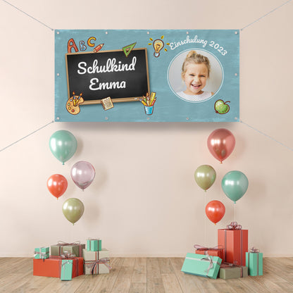 Personalisierbares Einschulungsbanner mit einer Kreidetafel, auf der "Schulkind Emma" steht, verziert mit Schulmotiven wie Bleistiften, Büchern und einem Apfel. Rechts oben befindet sich ein rundes Foto eines lachenden Mädchens sowie der Schriftzug "Einschulung 2023". Die Dekoration umfasst bunte Ballons und Geschenkpakete.