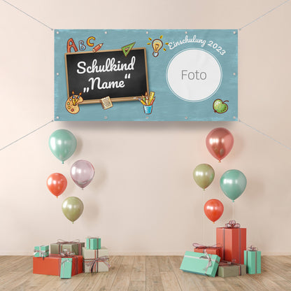 Personalisierbares Einschulungsbanner mit einer Kreidetafel, auf der "Schulkind 'Name'" steht, verziert mit Schulmotiven wie Bleistiften, einem Apfel und einer Glühbirne. Rechts oben ist ein runder Platzhalter für ein Foto mit der Aufschrift "Foto" sowie der Schriftzug "Einschulung 2023". Die Dekoration umfasst bunte Ballons und Geschenkpakete.