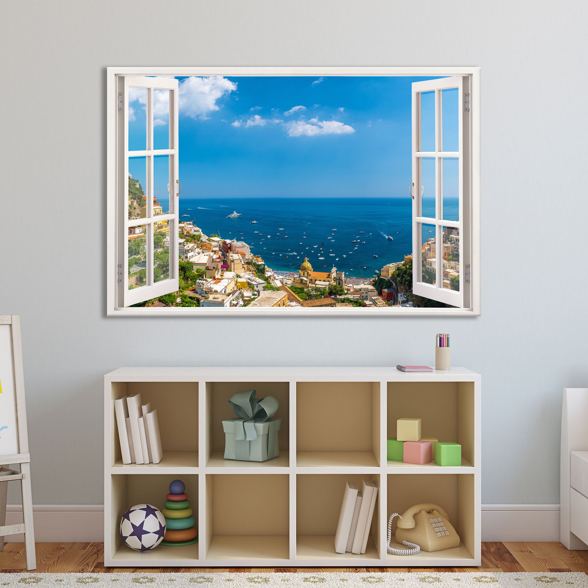 Wandtattoo mit einem offenen Fensterblick auf Positano an der Amalfiküste, perfekt für das Kinderzimmer oder den Wohnbereich. Die mediterrane Küstenlandschaft mit blauem Himmel, zahlreichen Booten und der malerischen Stadt verleiht dem Raum eine luftige und weite Atmosphäre. Das Motiv ist oberhalb eines weißen Regals mit Spielzeug, Büchern und Dekorationen angebracht und bringt eine entspannte Urlaubsstimmung in den Raum.