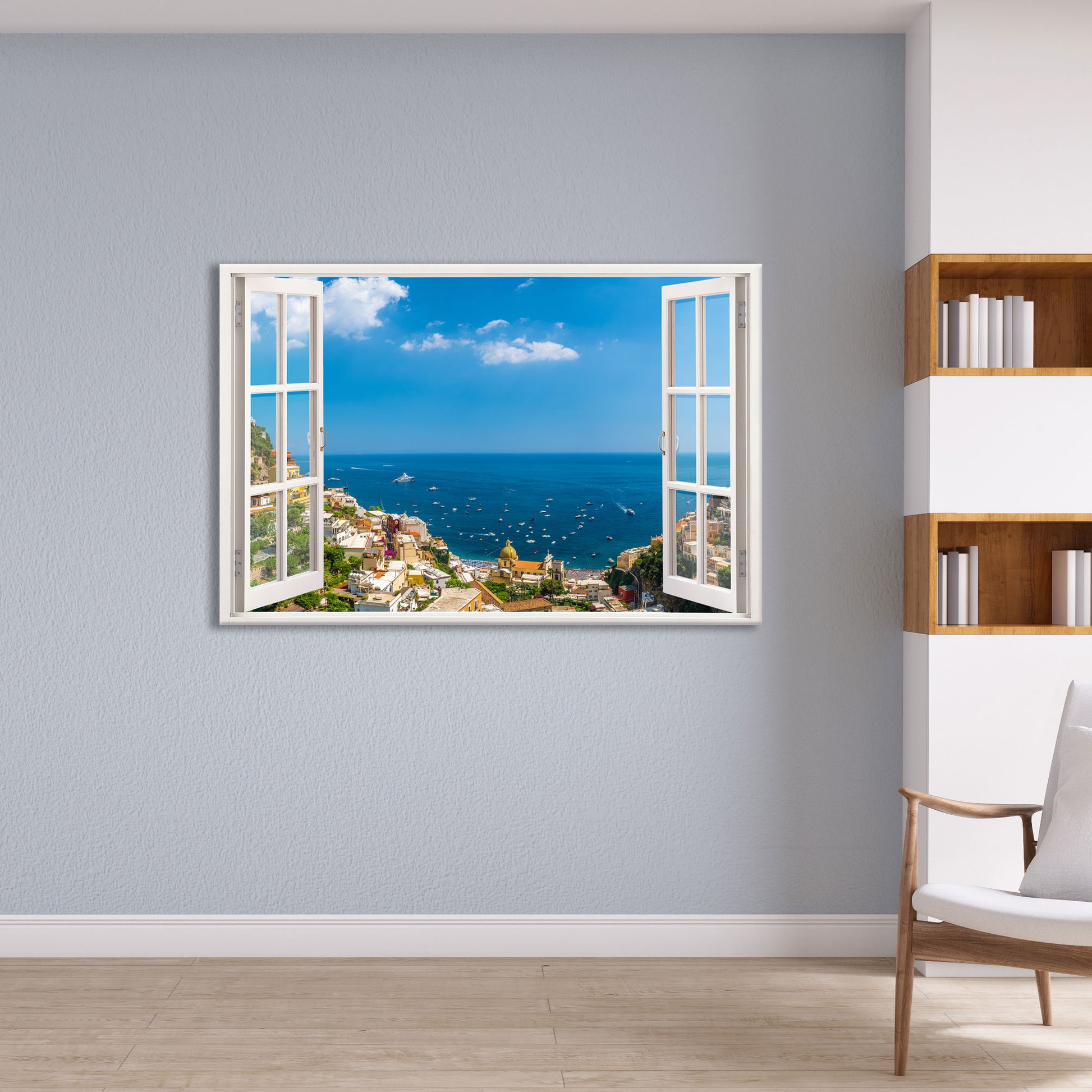 Ein Wandtattoo mit einem offenen Fensterblick auf Positano an der Amalfiküste, das eine malerische Aussicht auf das Mittelmeer bietet. Die helle, blaue Wand und das moderne Bücherregal mit Holz- und Weißelementen verleihen dem Raum eine stilvolle und elegante Atmosphäre. Das Fensterdesign mit weißen Rahmen erzeugt eine optische Illusion, als würde man direkt auf das Meer und die Küstenstadt blicken. Ideal für Wohnzimmer oder Arbeitsräume, um ein Gefühl von Weite und Urlaubsstimmung zu vermitteln.