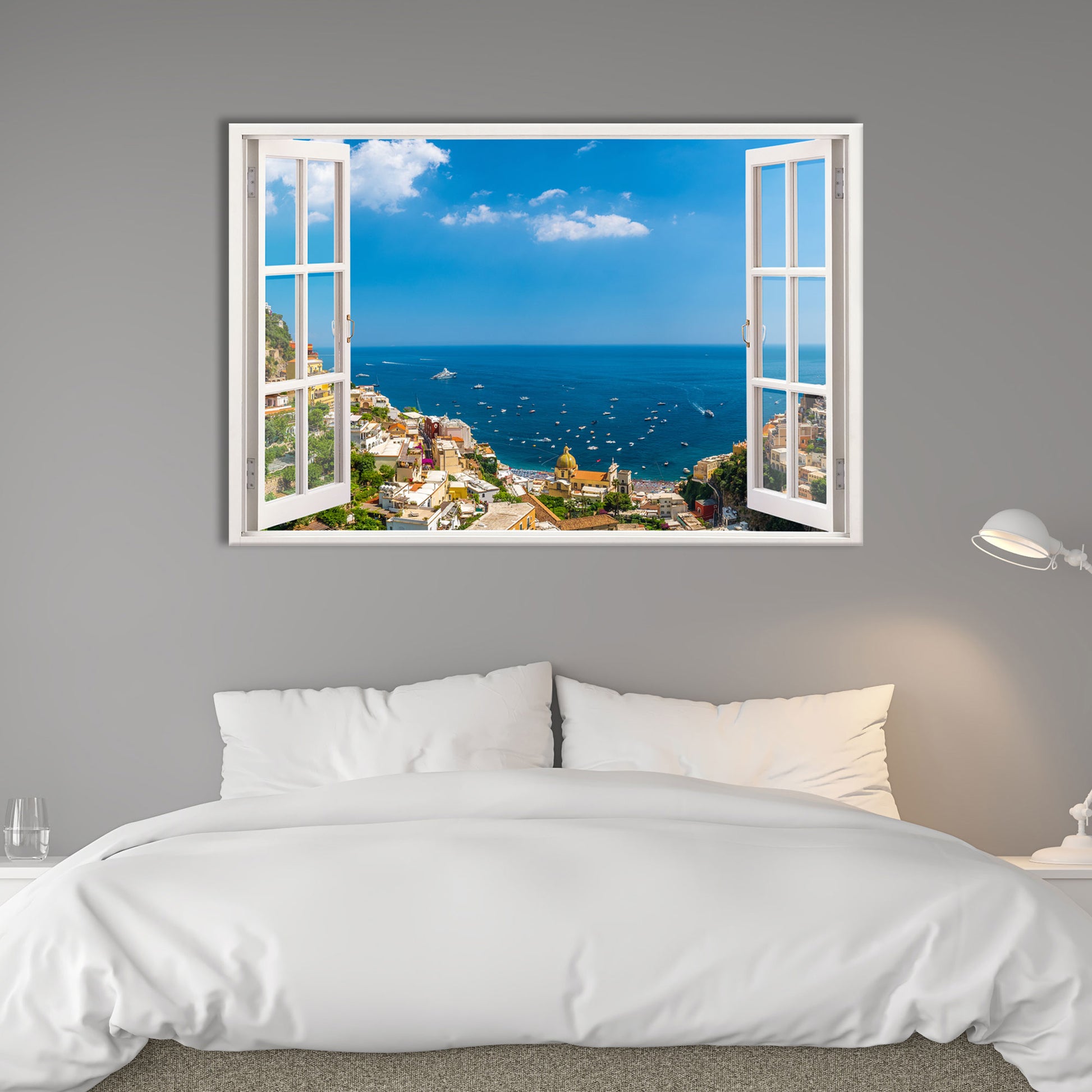 Ein beeindruckendes Wandtattoo mit einem offenen Fensterblick auf die italienische Küstenstadt Positano am Mittelmeer, das über einem gemütlichen Bett in einem modern eingerichteten Schlafzimmer angebracht ist. Die graue Wandfarbe und die weiße Bettwäsche sorgen für eine ruhige, harmonische Atmosphäre. Die stilvolle Nachttischlampe verleiht dem Raum ein gemütliches Ambiente. Das Fensterdesign mit weißen Rahmen erzeugt eine optische Täuschung, die den Raum größer erscheinen lässt.