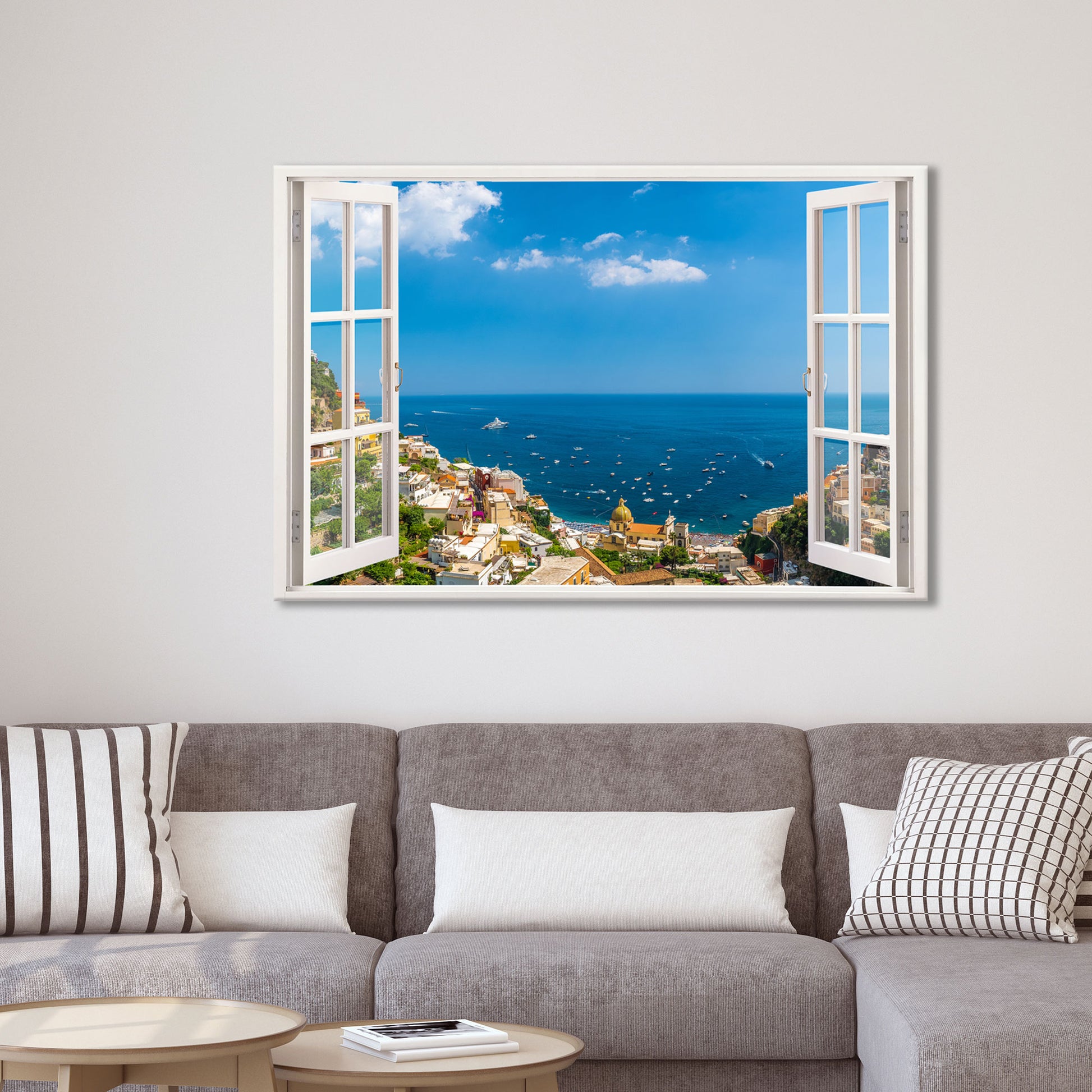 Ein dekoratives Wandtattoo mit einem offenen Fensterblick auf die malerische Küstenstadt Positano am Mittelmeer. Das Wandbild ist über einem modernen grauen Sofa mit weißen und gemusterten Kissen angebracht. Die realistische Fensteroptik verleiht dem Wohnzimmer eine offene, luftige Atmosphäre und erzeugt eine täuschend echte Aussicht auf das azurblaue Meer, die bunten Häuser und die idyllische Landschaft. Perfekt für eine entspannende und stilvolle Wohnraumgestaltung.