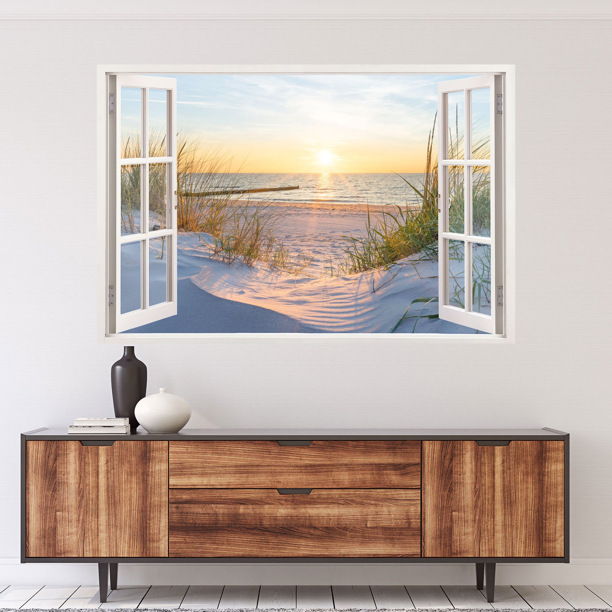 Wandtattoo mit offenem Fensterblick auf einen Sandstrand bei Sonnenuntergang. Die goldene Sonne spiegelt sich im ruhigen Meer, während sanfte Dünen mit Gräsern die Szene umrahmen. Perfekte Wanddekoration für eine entspannte, maritime Atmosphäre.